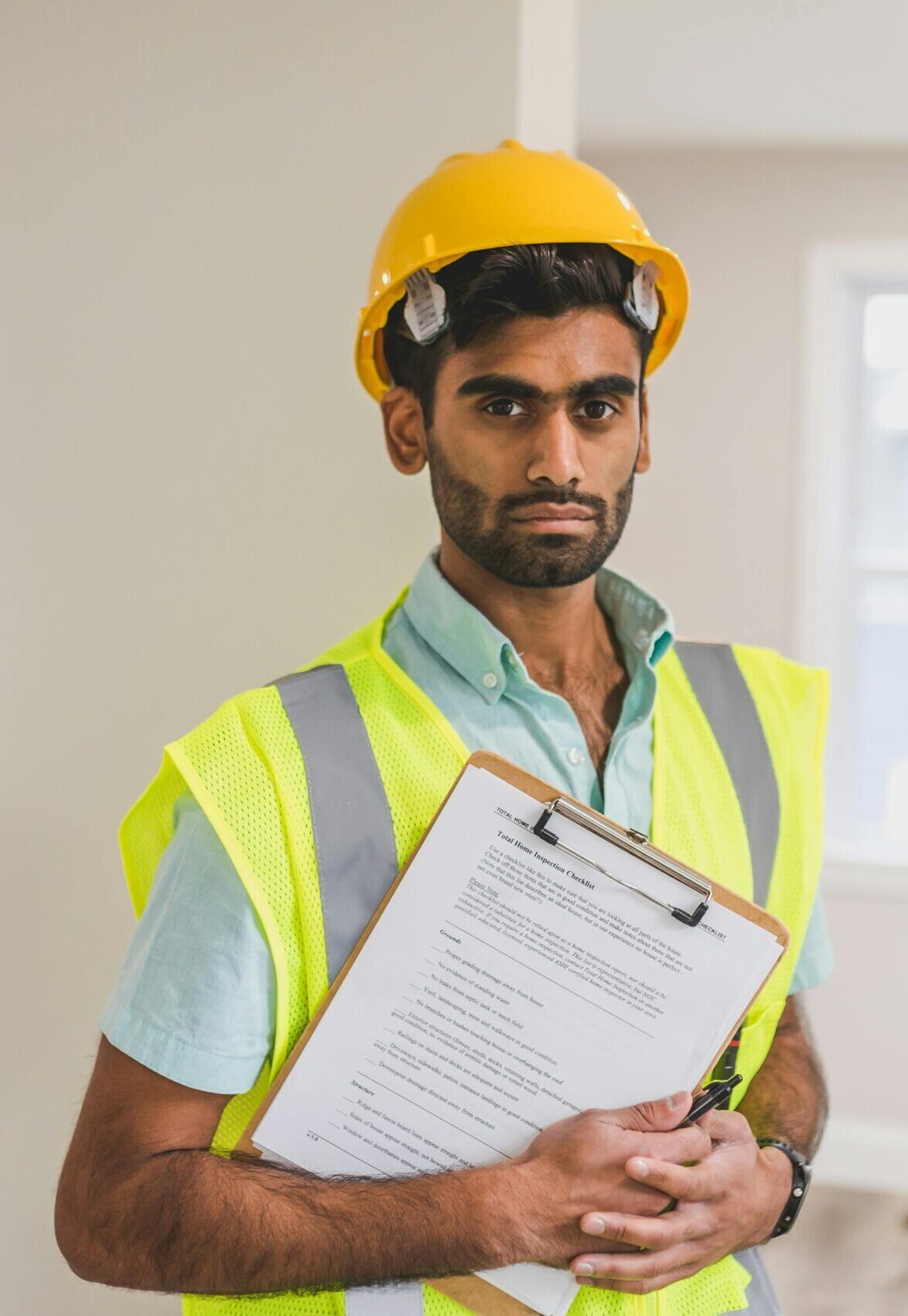 South Asian construction worker wearing safety gear and holding a clipboard indoors.
